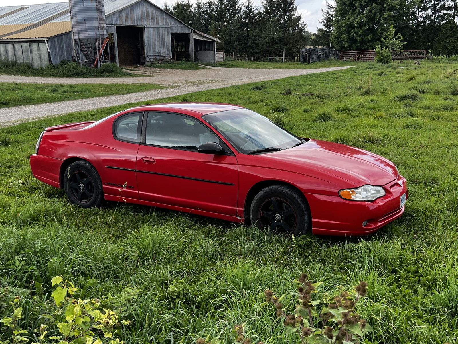 Red Chevrolet Monte Carlo parked in grass beside a barn.