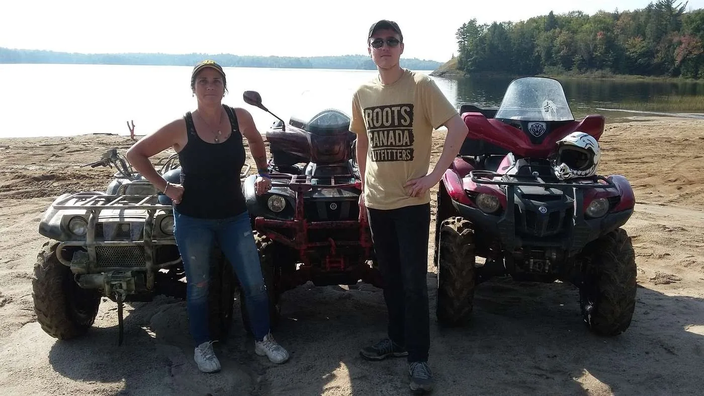 Two people standing beside all-terrain vehicles near a lake