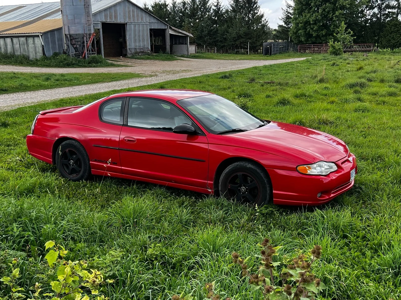 Red coupe parked in a grassy field beside a weathered barn
